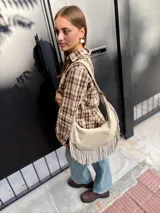 Cartera tipo hobo de cuero color arena, con diseño de media luna y flecos largos en la parte inferior. Cuenta con correa ajustable para llevar al hombro y cierre superior.