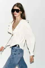 Blusa de georgette en color blanco, con escote en pico, detalle de volantes y lazada frontal. Presenta mangas largas abullonadas con puños ajustados y un bajo asimétrico con diseño tipo globo.