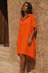 Vestido camisero corto de lino color naranja, con mangas largas arremangables mediante tira con botón, cierre frontal con botones y diseño amplio de corte asimétrico con la parte trasera más larga.