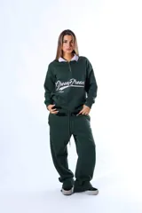 Polo de frisa color verde oscuro con cuello blanco y cierre parcial frontal. Presenta un bordado en el frente con el texto "Green Dreams" en tipografía cursiva blanca y la palabra "Aum" debajo.