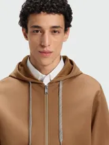 Buzo canguro con cierre frontal, color camel, con capucha y cordones trenzados. Presenta puños y ruedo con terminación de rib con franjas blancas y negras.