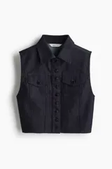 Chaleco corto de denim de algodón rígido, color azul marino, con cuello, cierre frontal de botones metálicos y bolsillos superiores con solapa y botón. Presenta un corte ajustado con costuras visibles para aportar estructura.