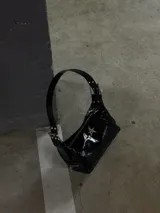 Bolso de hombro negro de charol, con forma alargada y cierre principal con cremallera. Presenta un dije metálico con forma de estrella de cuatro puntas en el frente y detalles de cremalleras decorativas en los laterales.