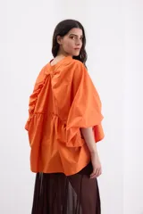 Camisa de corte holgado y diseño oversize, color naranja vibrante. Presenta cuello redondo con detalle de volado, cierre frontal con botones y mangas anchas con caída de hombro. Confeccionada en algodón con textura suave.