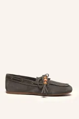 Mocasín tipo náutico confeccionado en gamuza gris, con cordones frontales decorados con borlas y cuentas de colores. Cuenta con suela plana de goma.