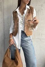 Chaleco de gamuza color camel con bordados en hilo blanco y detalles de tachas metálicas. Presenta un diseño abierto con terminaciones redondeadas en el frente.