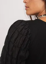 Blusa negra de tejido de punto acanalado, con cuello redondo y mangas largas abullonadas de tela semitransparente. Presenta una hilera vertical de pequeñas cuentas plateadas en el frente.