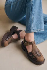Sandalia tipo cangrejera de cuero lavado en tonos marrones, con diseño de tiras entrelazadas, puntera cerrada y suela de goma dentada tipo track. Cuenta con ajuste mediante correa con hebilla metálica en el tobillo.