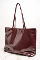 Bolso tipo tote de gran tamaño en color bordó con acabado brillante. Cuenta con doble asa de hombro y placa metálica decorativa con logo en la parte frontal.