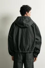 Cazadora de denim color negro con capucha ajustable con cordones, cierre frontal de cremallera, bolsillos tipo canguro y detalles de costuras en contraste en las mangas.