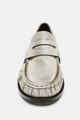 Mocasín de piel color gris claro con efecto desgastado, detalle de fruncidos en la puntera y antifaz en el empeine. Posee un taco bajo y suela oscura.