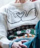 Sweater artesanal de lana merino en color natural, con diseño tejido de jacquard en la parte inferior y bordado a mano de dos cisnes enfrentados formando un corazón en el pecho. Presenta cuello redondo, puños y ruedo en tono verde contrastante.