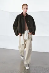 Chaqueta encerada de corte oversize, color marrón oscuro, con cuello de pana en contraste. Presenta cierre frontal con cremallera y botones a presión ocultos bajo solapa, bolsillos delanteros y mangas largas con puños ajustables.