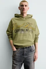 Sudadera con capucha de corte relajado en color verde oliva con efecto lavado. Presenta un bolsillo frontal tipo canguro y un estampado gráfico en el pecho con letras en tonos amarillos y negros. Cuenta con puños y bajo acanalados.