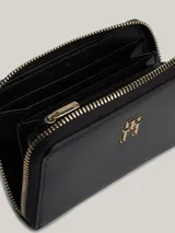 Billetera negra de poliuretano con cierre de cremallera y logo metálico dorado de Tommy Hilfiger en el frente.