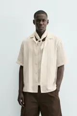 Camisa de calce relajado confeccionada en tejido con algodón de color beige. Presenta cuello bowling, manga corta, aberturas laterales en el bajo y cierre frontal con botones.