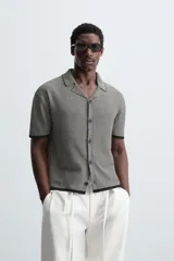 Camisa de punto de manga corta con cuello bowling y cierre frontal de botonadura. Presenta un patrón de rayas horizontales finas en blanco y negro, con ribetes negros en cuello, puños y bajo.