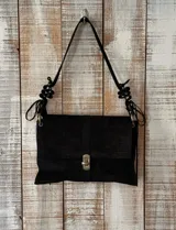 Cartera tipo bolso de mano color negro con textura de cuero croco. Presenta un cierre frontal metálico y una correa ajustable con detalles decorativos de nudos en los extremos.