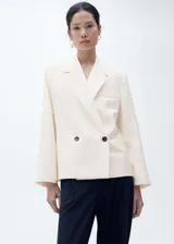 Blazer color crema de corte recto con cierre cruzado de doble botonadura, cuello con solapa de muesca, manga larga con puños abotonados, dos bolsillos de ribete frontales y botones en contraste.