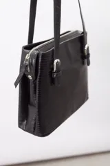 Cartera de hombro estructurada color negro, confeccionada en 100% cuero vacuno con textura en relieve tipo reptil. Cuenta con asas largas ajustables mediante hebillas metálicas plateadas, cierre superior y el logo de la marca grabado en la parte frontal.