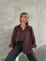 Camisa de corte holgado color marrón, con cuello clásico, cierre frontal de botones y detalle de bordado en la zona de los hombros y canesú. Presenta mangas largas con volumen.