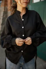 Camisa de lino color negro con silueta relajada, cuello camisero y cierre frontal con botones de nácar. Presenta mangas largas con puños y detalles de tiras laterales.