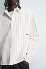 Camisa de corte relajado confeccionada en tejido de algodón, con diseño de rayas verticales. Presenta cuello solapa, cierre frontal de botones, manga larga con puños abotonados y un bolsillo plastrón en el pecho con un pequeño bordado decorativo.