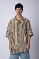Camisa de manga corta con cuello solapa, corte holgado y cierre frontal de botones. Presenta un diseño de rayas verticales en tonos beige, negro y blanco.