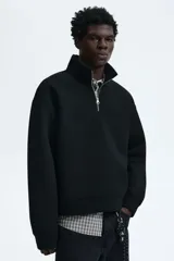 Buzo negro de tejido pesado con efecto neopreno, cuello elevado y medio cierre frontal plateado. Presenta hombros caídos, mangas largas y puños y bajo acanalados.