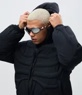 Campera puffer negra con diseño acolchado que simula una estructura de esqueleto. Cuenta con capucha, cierre frontal con cremallera, mangas largas y puños elásticos.