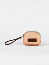 Monedero de cuero color salmón con cierre superior y correa de muñeca de cuero marrón. Presenta un parche decorativo con el logo de la marca en la parte frontal.