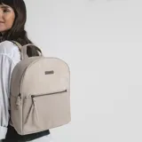 Mochila color beige con aplique de cuero sintético en la parte inferior y superior. Tiene un bolsillo frontal con cierre y un bolsillo interno. Las correas son regulables.